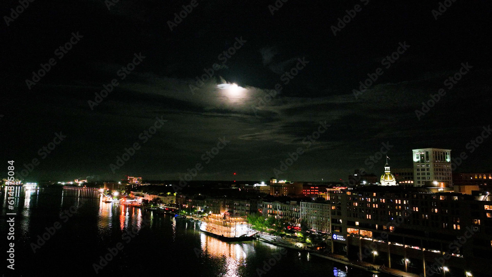 Drone photo at night over the Savannah River. Flying over ferry boat a brightly lit boardwalk with moon in distance.