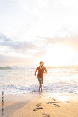 Vertical image of a smiling young man running through the waves at the shore of the beach on a summer day with beautiful sunset light.