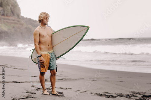 Fit young surfer man with curly blond hair with surfboard goes by the ocean having fun doing extreme water sports, surfing. Travel and healthy lifestyle concept. Sports travel destination.