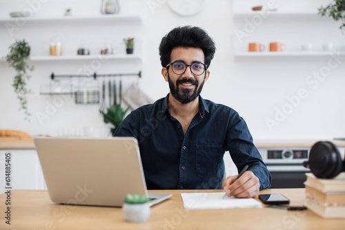 Portrait of attractive indian businessman posing at office desk with digital devices in spacious dining room. Self-employed worker carrying out professional activity in comfortable workplace.