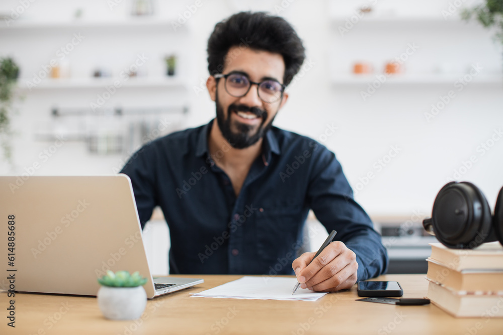 Portrait of attractive indian businessman posing at office desk with digital devices in spacious dining room. Self-employed worker carrying out professional activity in comfortable workplace. Portrait of attractive indian businessman posing at office desk with digital devices in spacious dining room. Self-employed worker carrying out professional activity in comfortable workplace.