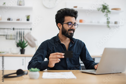 Attractive bearded man in spectacles holding blank credit card while typing on portable computer in kitchen interior. Happy arabian gentleman doing online banking transaction while working remotely.