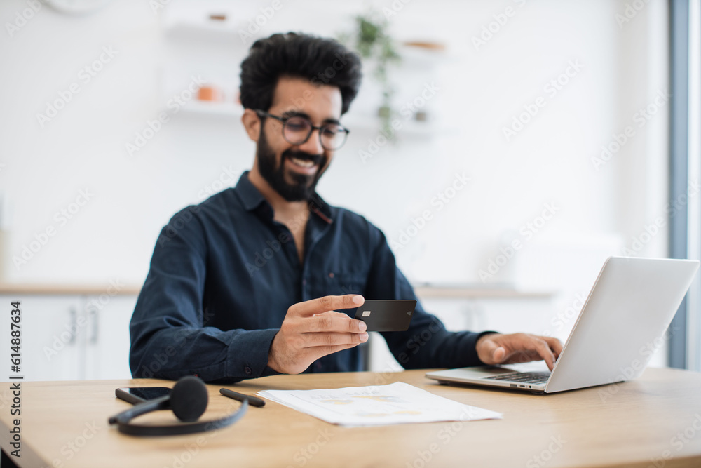 Fototapeta premium Joyful arabian man in casual clothes using black credit card while running portable computer in workplace. Smiling bearded person in glasses enjoying virtual shopping using online wallet from home.