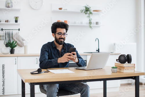 Positive indian male in spectacles holding smartphone while staying in modern workplace at home. Efficient freelance worker texting message to colleague while performing project for employer indoors.