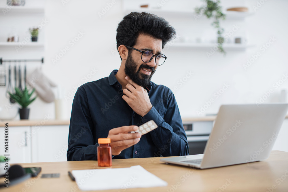 Feverish bearded man in casual clothes touching forehead while looking at laptop camera in dining room of apartment. Unwell arabian manager arranging virtual appointment with health professional. Feverish bearded man in casual clothes touching forehead while looking at laptop camera in dining room of apartment. Unwell arabian manager arranging virtual appointment with health professional.