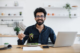 Portrait of arabian gentleman posing with vegetarian salad near computer at writing desk in home office. Cheerful business person increasing energy while choosing healthy diet in distant workplace.
