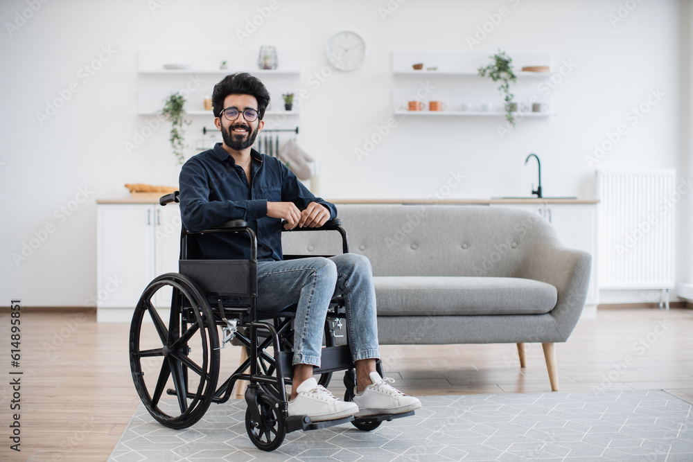 Full length portrait of arabian wheelchair user smiling at camera while resting in open-plan kitchen of modern apartment. Cheerful young male in casual clothes and glasses spending pastime indoors. Full length portrait of arabian wheelchair user smiling at camera while resting in open-plan kitchen of modern apartment. Cheerful young male in casual clothes and glasses spending pastime indoors.