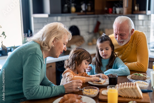 Happy seniors male and female having breakfast with two small children at dinning table in the kitchen at home.