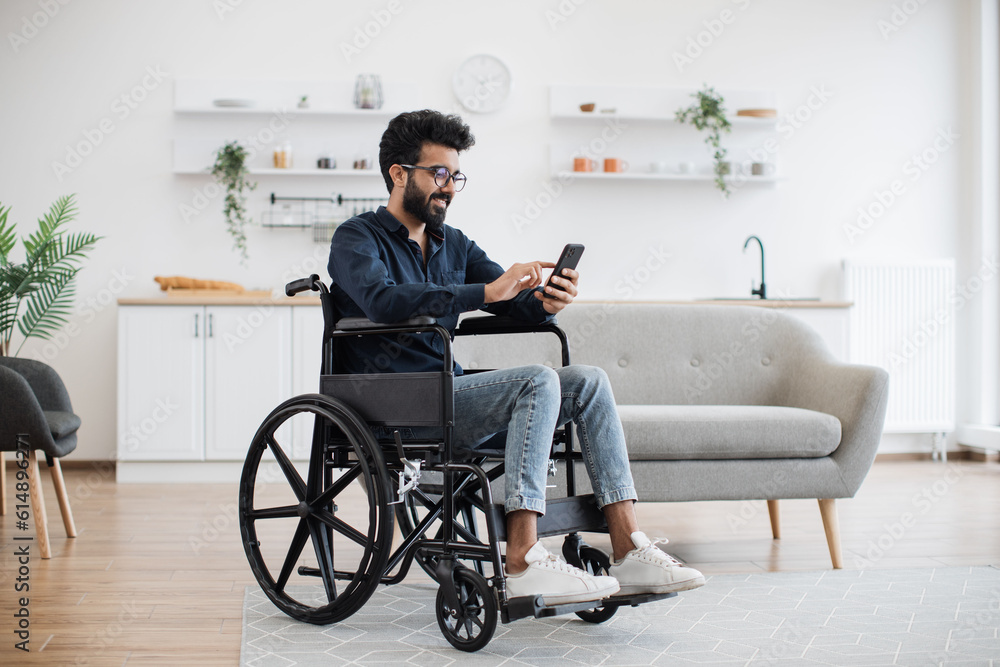 Full length view of indian male with disability using smartphone while resting in modern kitchen of apartment. Joyful person in glasses and casual clothes texting message on digital device at home. Full length view of indian male with disability using smartphone while resting in modern kitchen of apartment. Joyful person in glasses and casual clothes texting message on digital device at home.