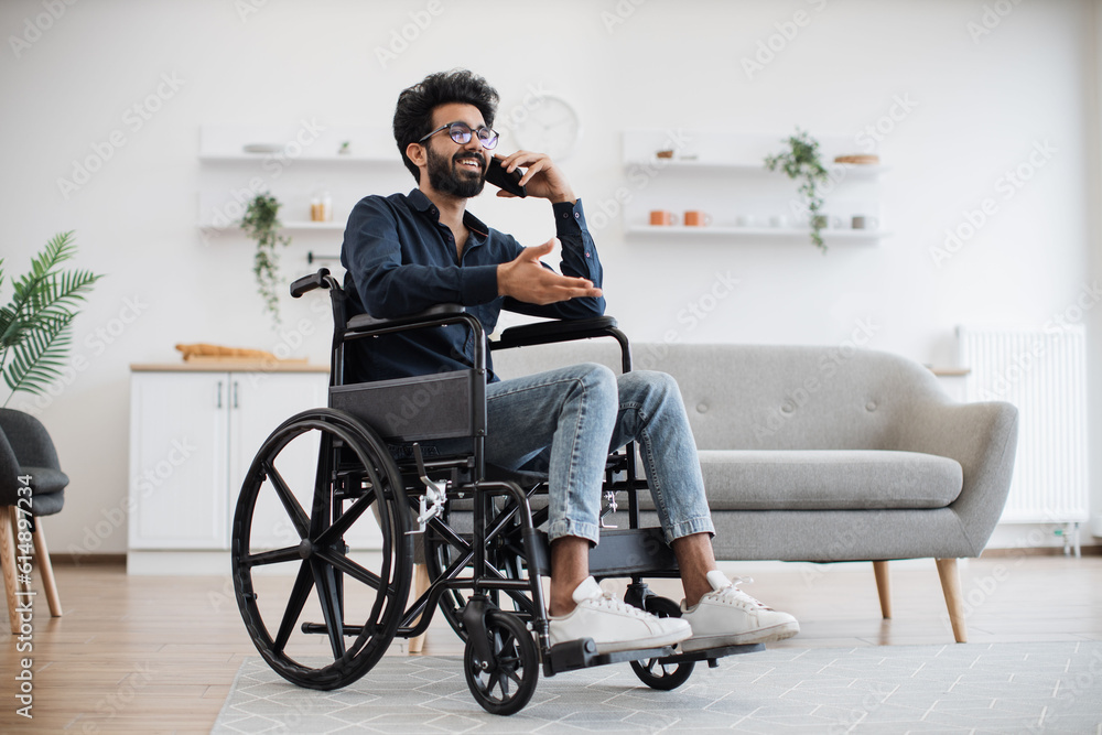 Full length view of adult person in wheelchair talking on cell phone while using comfort of modern studio flat. Mature arabian man organising birthday party while getting recovery from illness. Full length view of adult person in wheelchair talking on cell phone while using comfort of modern studio flat. Mature arabian man organising birthday party while getting recovery from illness.