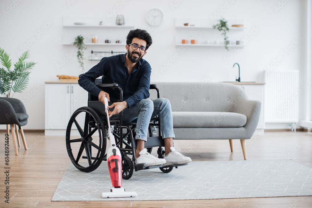 Helpful young man with disability holding cordless vacuum cleaner while sitting in wheelchair in open-plan kitchen. Active person in casual outfit collecting dust and dirt from carpet in apartment. Helpful young man with disability holding cordless vacuum cleaner while sitting in wheelchair in open-plan kitchen. Active person in casual outfit collecting dust and dirt from carpet in apartment.