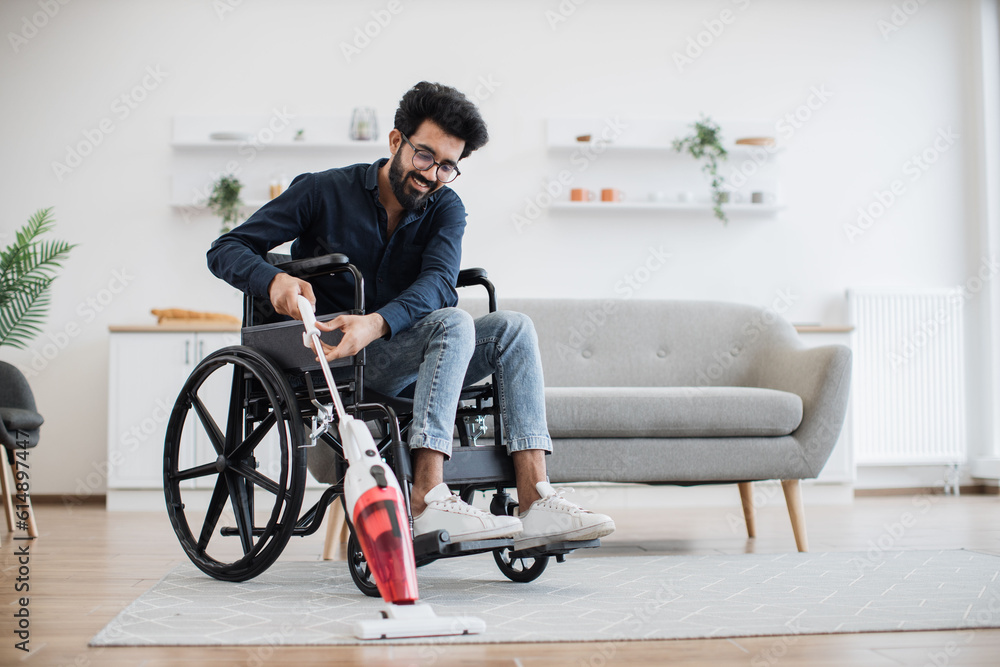 Relaxed adult person in jeans taking lightweight stick vacuum while using wheelchair in spacious flat. Cheerful indian homeowner enjoying housework with modern appliances while waiting for guests. Relaxed adult person in jeans taking lightweight stick vacuum while using wheelchair in spacious flat. Cheerful indian homeowner enjoying housework with modern appliances while waiting for guests.