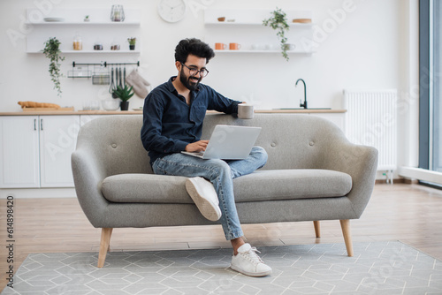 Full length view of handsome bearded man with cup of tea in hand using modern laptop while resting on sofa at home. Young arabian person in glasses surfing internet webpages at leisure time indoors.