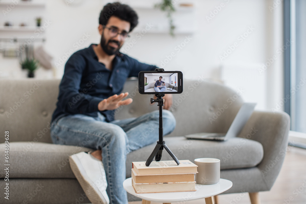 Positive indian influencer with black beard recording video on modern smartphone while sitting on couch with laptop. Happy young man sharing his success with subscribers online.