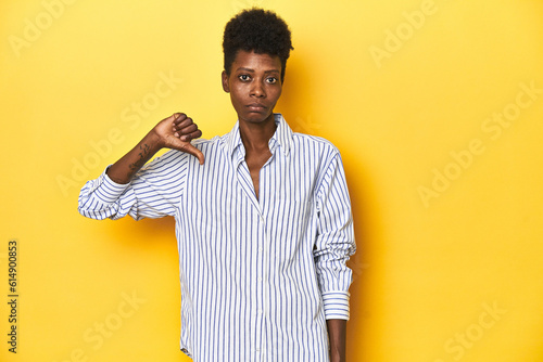 African businesswoman  blue striped shirt  yellow backdrop  showing a dislike gesture  thumbs down. Disagreement concept.