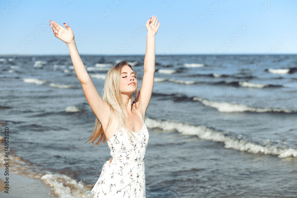 Happy blonde beautiful woman on the ocean beach standing in a white summer dress, raising hands Happy blonde beautiful woman on the ocean beach standing in a white summer dress, raising hands