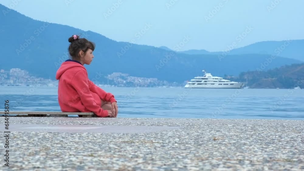 Sad child on the sea embankment. A lonely little girl sits on the embankment against the backdrop of a luxury yacht. Sad child on the sea embankment. A lonely little girl sits on the embankment against the backdrop of a luxury yacht.