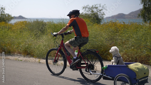 Elderly senior man biking on an electric bike on a trail pulling a trailer with a dog in it.
