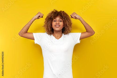 young curly american girl with braces shows biceps over yellow isolated background