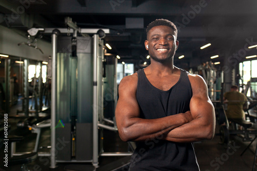young African American man stands with crossed arms in dark gym and smiles, fitness trainer stands in fitness club