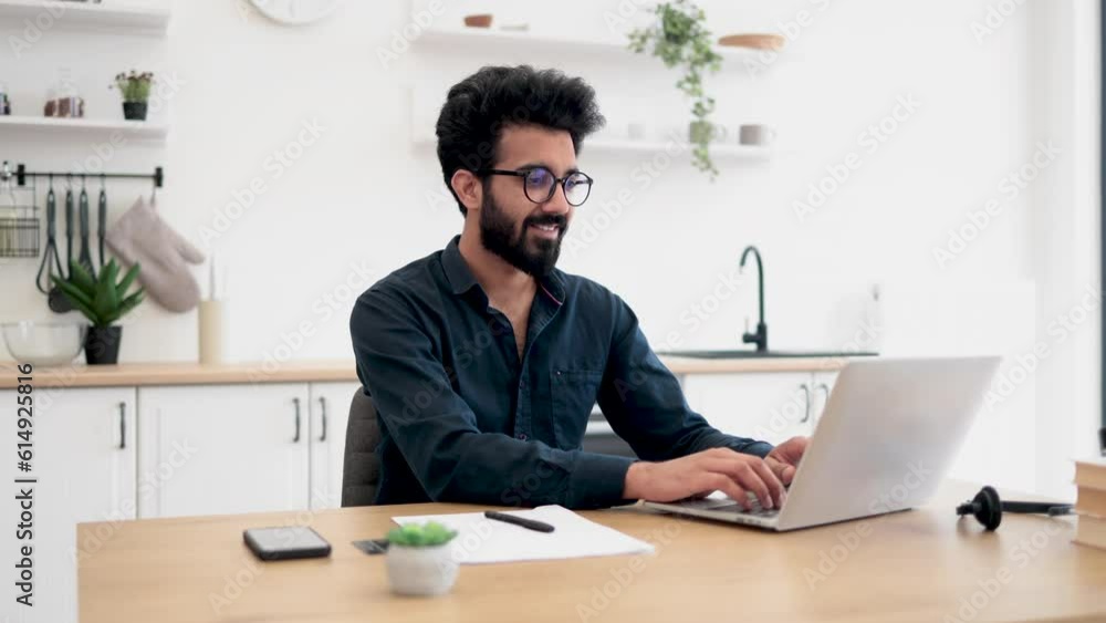 Joyful bearded man in glasses operating modern laptop while sitting at writing desk in spacious dining room. Smiling indian freelancer using flexible schedule while spending working hours at home. Joyful bearded man in glasses operating modern laptop while sitting at writing desk in spacious dining room. Smiling indian freelancer using flexible schedule while spending working hours at home.