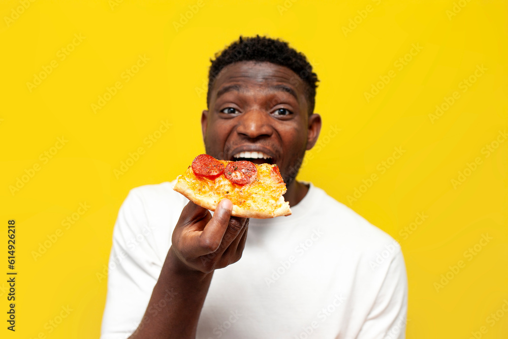 joyful african american man in white t-shirt bites piece of pizza on yellow isolated background, guy eats fast food