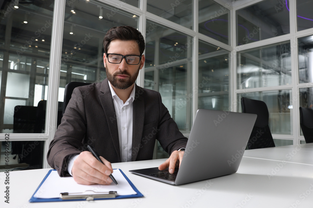 Man writing notes while working on laptop at white desk in office Man writing notes while working on laptop at white desk in office