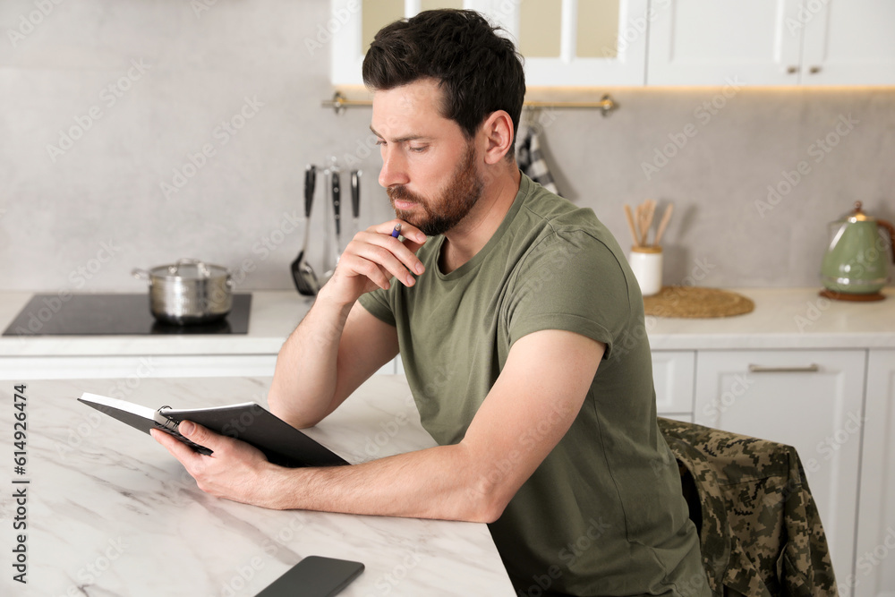 Soldier with notebook at white marble table in kitchen. Military service Soldier with notebook at white marble table in kitchen. Military service
