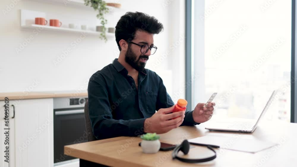 Anxious adult person holding round pills while having online conversation via computer on kitchen background. Aching indian man receiving doctor's consultation via internet connection at home. Anxious adult person holding round pills while having online conversation via computer on kitchen background. Aching indian man receiving doctor's consultation via internet connection at home.