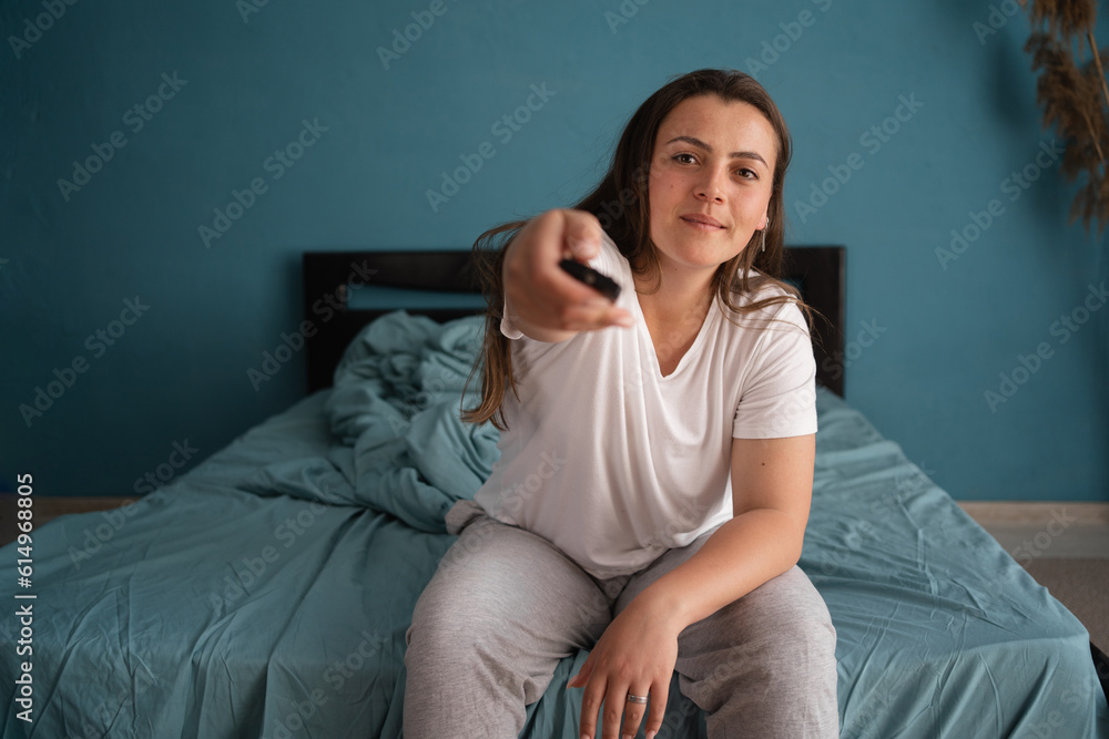 Woman watches TV sitting on the bed, using tv remote control in her hands. Morning or evening in the bedroom Woman watches TV sitting on the bed, using tv remote control in her hands. Morning or evening in the bedroom