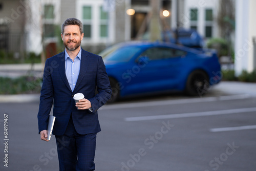 Businessman walking in city. Gray hair business man walk on city street. Business man walking outdoor, hold laptop take away coffee cup. Business man in suit go to office work. Successful business. © Volodymyr