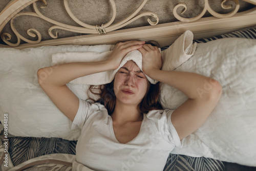 Young woman with headache flu ill sick disease cold at home indoor lying on bed with cold compress on her head at bedroom home