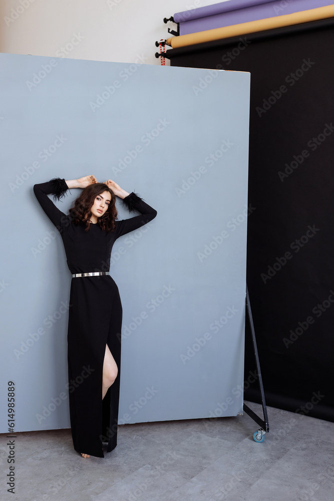 Attractive woman with long brunette hair, dressed in a trendy black long-sleeved dress, posing, looking away. A female model standing against a black and light blue wall in the background.
