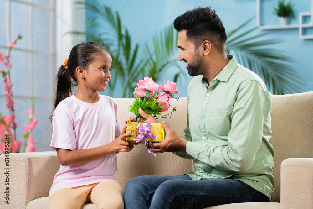 Happy young Indian girl kid giving flowers with gift to father while sitting on sofa at home - concept of father's day, relationship and family affection. Happy young Indian girl kid giving flowers with gift to father while sitting on sofa at home - concept of father's day, relationship and family affection.
