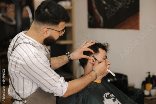 Side view of barber using sharp blade for shaving. Handsome customer sitting in barbershop