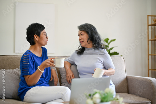Two mature middle-aged Asian women are enjoying their morning coffee and talking on a sofa