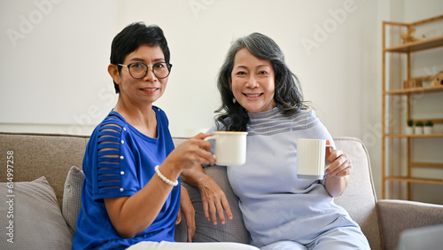 Two retired Asian women are sitting on a sofa in the living room with a coffee cup in their hands.