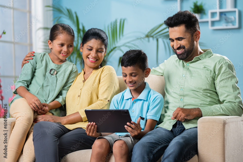 Happy Indian parents sitting with sibling kids while son using digital tablet at home - concept of family bonding or time and weekend holidays and cyberspace Happy Indian parents sitting with sibling kids while son using digital tablet at home - concept of family bonding or time and weekend holidays and cyberspace