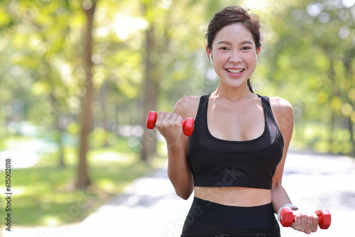 Beautiful asian fitness female sportswoman lifting dumbbell weight training after finish morning run outdoor. Happy smiling athlete woman excercising under the tree in the park while staring at camera