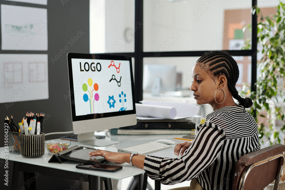 Side view of young serious African American female webdesigner sitting by workplace in front of computer monitor and using tablet Side view of young serious African American female webdesigner sitting by workplace in front of computer monitor and using tablet
