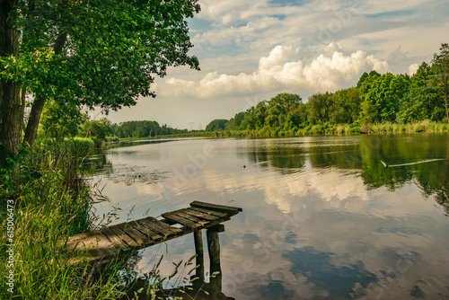 Widok na stary drewniany pomost na jeziorze, chmurki odbijające się w wodzie, zieleń drzew