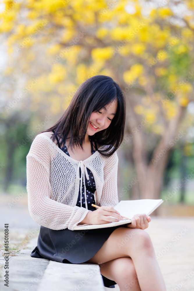 Portrait cute smiles Asian of attractive young teenage girl a holding diary book writing note at Yellow Tabebuia Chrysotricha flowers with the park in spring day at Evening background in Thailand. Portrait cute smiles Asian of attractive young teenage girl a holding diary book writing note at Yellow Tabebuia Chrysotricha flowers with the park in spring day at Evening background in Thailand.
