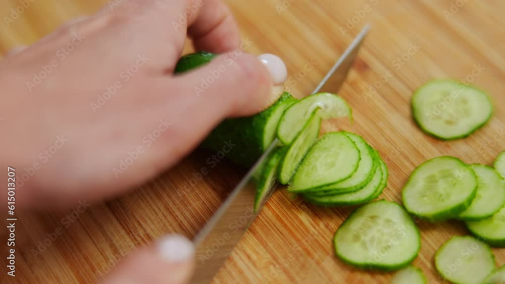 High angle view close-up female hand slicing cucumber with knife on cutting board. Closeup unrecognizable young Caucasian woman cutting ingredient for salad indoors High angle view close-up female hand slicing cucumber with knife on cutting board. Closeup unrecognizable young Caucasian woman cutting ingredient for salad indoors