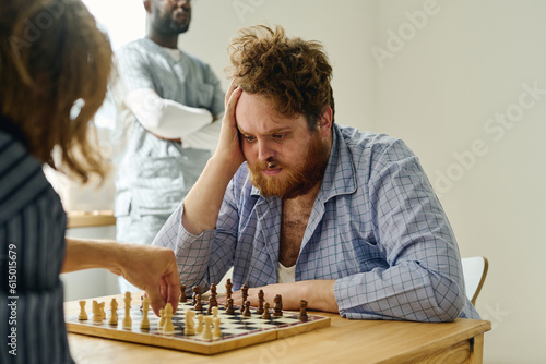 Nervous man with pawn in his nose looking at chess game partner making move while both sitting in front of each other by wooden table