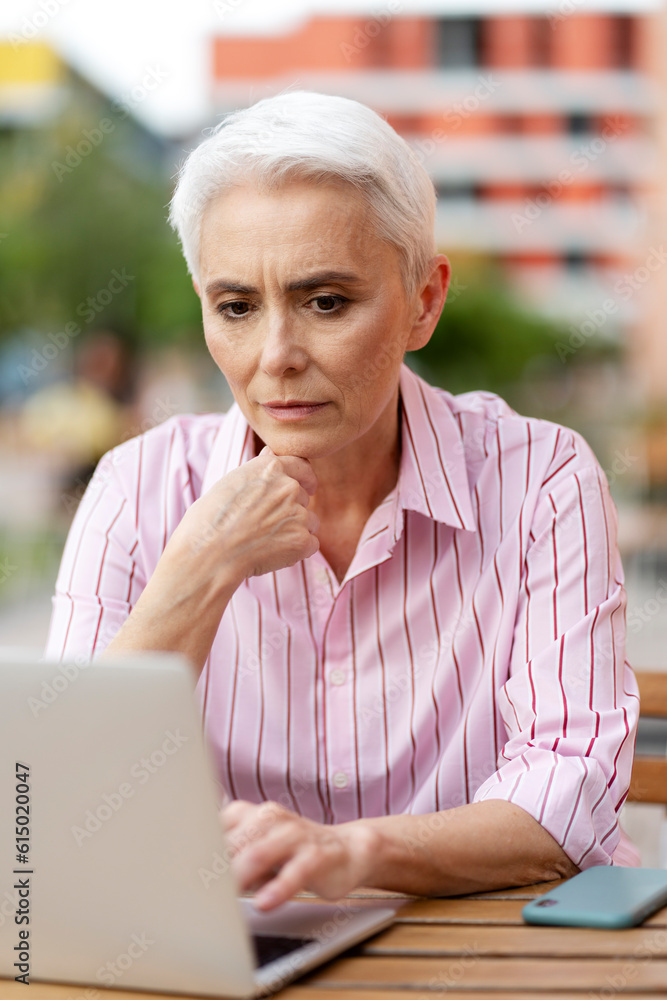 Pensive elderly woman working at computer, sitting at workplace, online work on street. Attractive freelancer working by workplace. Successful business concept Pensive elderly woman working at computer, sitting at workplace, online work on street. Attractive freelancer working by workplace. Successful business concept