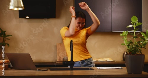 Smiling young woman working on a laptop and dancing in her kitchen at home