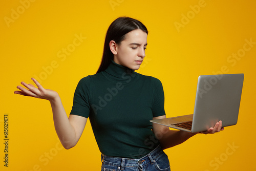 Young caucasian woman with a laptop having doubts while raising a hand, isolated over yellow background.