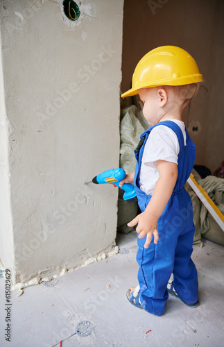 Child drilling wall with toy electric drill tool in apartment under renovation. Kid in work overalls using drilling construction instrument at home during refurbishment.