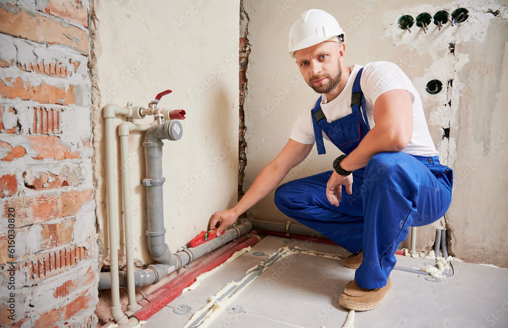 Male plumber using spirit level tool while installing pipe system at home, looking to the camera. Man measuring pipe level with building instrument in apartment under renovation.