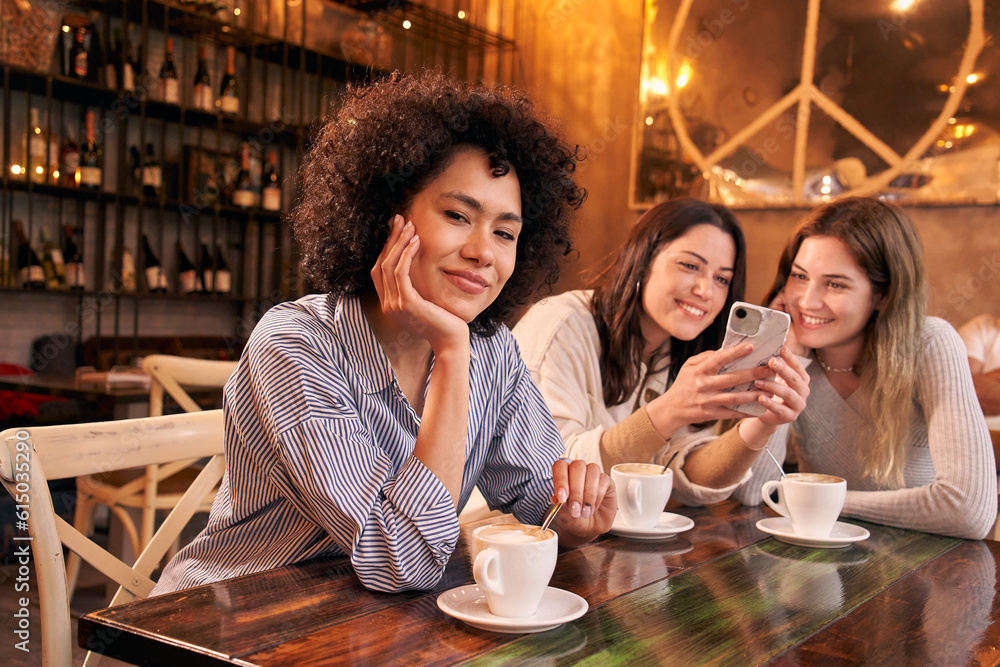 Portrait of a happy Latin woman in a cafe bar looking at the camera. People indoors staring at a smartphone at the same table while having coffee. Female girlfriends spending time together